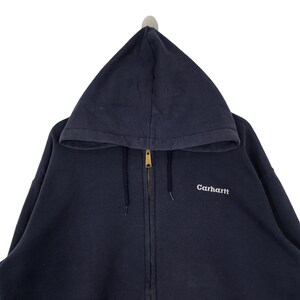 K&ouml;nnte beinhalten: Ein marineblauer Kapuzenpullover mit Rei&szlig;verschluss und Kordelzug. Der Hoodie hat einen goldfarbenen Rei&szlig;verschluss und den wei&szlig; gestickten Markennamen "Carhartt" auf der Brust. Das Kleidungsst&uuml;ck h&auml;ngt vor wei&szlig;em Hintergrund.