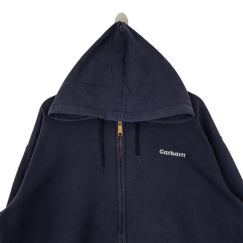 K&ouml;nnte beinhalten: Ein marineblauer Kapuzenpullover mit Rei&szlig;verschluss und Kordelzug. Der Hoodie hat einen goldfarbenen Rei&szlig;verschluss und den wei&szlig; gestickten Markennamen "Carhartt" auf der Brust. Das Kleidungsst&uuml;ck h&auml;ngt vor wei&szlig;em Hintergrund.