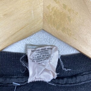 Puede incluir: Primer plano de una prenda negra con una etiqueta blanca. La etiqueta tiene instrucciones de cuidado en texto negro: "MACHINE WASH WARM, TUMBLE DRY, LOW SETTING, DO NOT BLEACH, DECORATED DO NOT DRY CLEAN OR IRON, RN 46831."