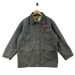 May include: A grey field jacket featuring a tan sherpa lining, a fold-down collar, and four front pockets with flaps. A rectangular patch on the upper left chest displays the text "MAGICAL MUSHROOMS MMM" in black letters over a yellow and black striped background.