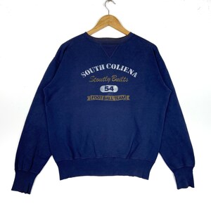 May include: Navy blue sweatshirt featuring the text "SOUTH COLIENA Stoutly Built" and "54 FOOT BALL TEAM" in a vintage style. The long-sleeved crew neck top is hanging on a wooden hanger.