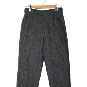 K&ouml;nnte beinhalten: Eine graue Hose mit hohem Bund, Knopfverschluss und Falten. Die Hose h&auml;ngt an einem Holzb&uuml;gel. Der Stoff hat ein strukturiertes Aussehen.