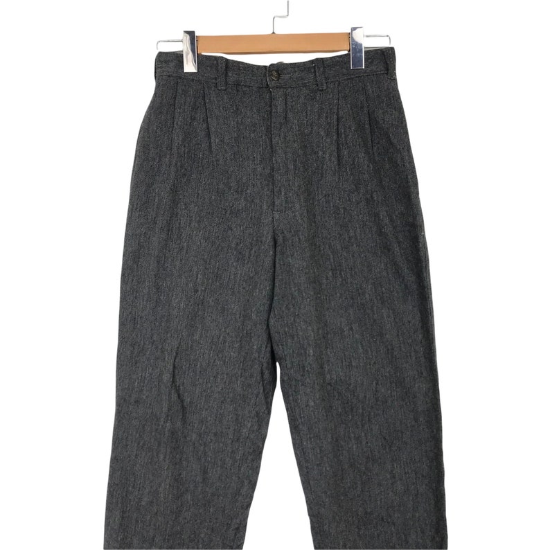 K&ouml;nnte beinhalten: Eine graue Hose mit hohem Bund, Knopfverschluss und Falten. Die Hose h&auml;ngt an einem Holzb&uuml;gel. Der Stoff hat ein strukturiertes Aussehen.