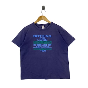 Könnte beinhalten: Ein marineblaues T-Shirt mit dem Text "NOTHING TO LOSE EVOLUTION IS THE ACT OF POSITIVE CHANGE PRODUCE DEVEROPMENT PEARL DIVER 1999" in Blau und Grün. Das T-Shirt hängt an einem Holzkleiderbügel.