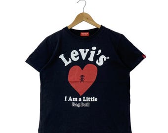 May include: A black short-sleeve t-shirt featuring the white text Levi's above a large red heart graphic with a small doll illustration inside. Below the heart, the white text reads I Am a Little Rag Doll. A small red brand tag is on the left sleeve.