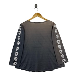 Puede incluir: Una camiseta de manga larga con un degradado de color gris oscuro a negro. Las mangas est&aacute;n adornadas con un patr&oacute;n repetitivo de calaveras blancas impresas a lo largo de cada brazo. La prenda tiene un cuello redondo y cuelga de una percha.