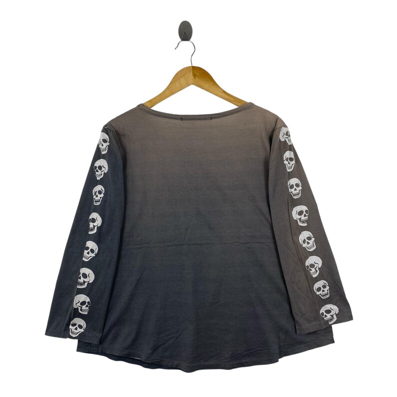 Puede incluir: Una camiseta de manga larga con un degradado de color gris oscuro a negro. Las mangas est&aacute;n adornadas con un patr&oacute;n repetitivo de calaveras blancas impresas a lo largo de cada brazo. La prenda tiene un cuello redondo y cuelga de una percha.