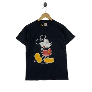 Puede incluir: Camiseta negra con un gr&aacute;fico vintage de Mickey Mouse. El personaje de dibujos animados se muestra en su pose cl&aacute;sica, con detalles en blanco, rojo y amarillo. La camiseta est&aacute; colgada en una percha de madera.