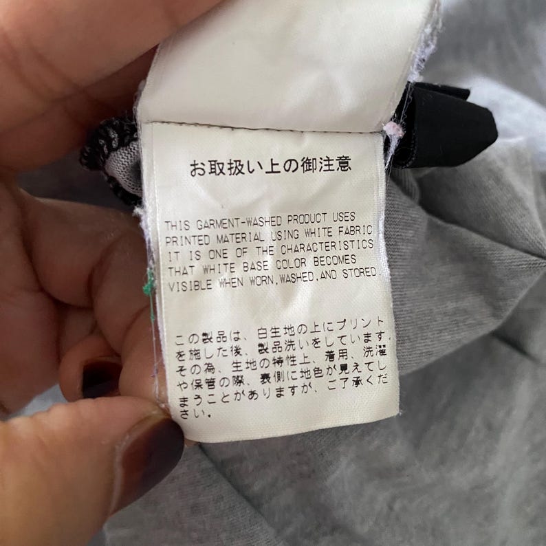 Puede incluir: Una etiqueta de cuidado rectangular blanca con texto negro en ingl&eacute;s y japon&eacute;s. El texto en ingl&eacute;s dice: THIS GARMENT-WASHED PRODUCT USES PRINTED MATERIAL USING WHITE FABRIC. IT IS ONE OF THE CHARACTERISTICS THAT WHITE BASE COLOR BECOMES VISIBLE WHEN WORN, WASHED, AND STORED.