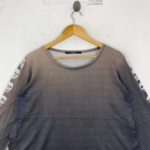 Puede incluir: Una camiseta de manga larga con un efecto degradado de marr&oacute;n a gris oscuro. Las mangas est&aacute;n decoradas con un patr&oacute;n repetitivo de calaveras blancas. La prenda tiene un cuello redondo y una etiqueta interior negra con el texto LIMI feu.