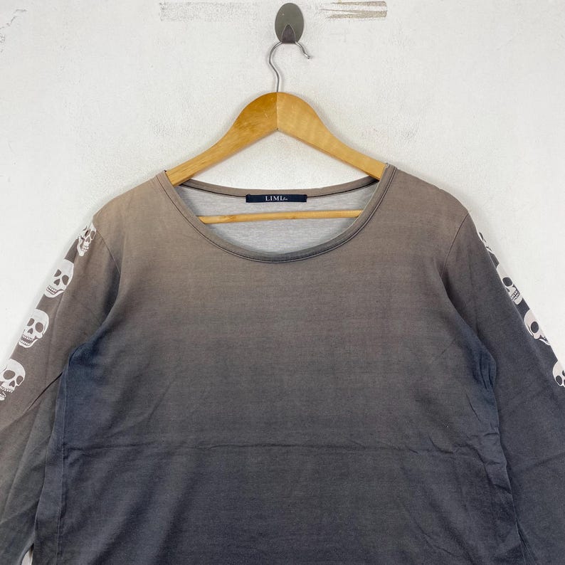 Puede incluir: Una camiseta de manga larga con un efecto degradado de marr&oacute;n a gris oscuro. Las mangas est&aacute;n decoradas con un patr&oacute;n repetitivo de calaveras blancas. La prenda tiene un cuello redondo y una etiqueta interior negra con el texto LIMI feu.