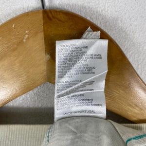 May include: A white fabric garment care label attached to a wooden hanger. The label features printed text in multiple languages including 100% Cotton, machine wash instructions at 30C/86F, and Made in Portugal. The tag is rectangular and white.