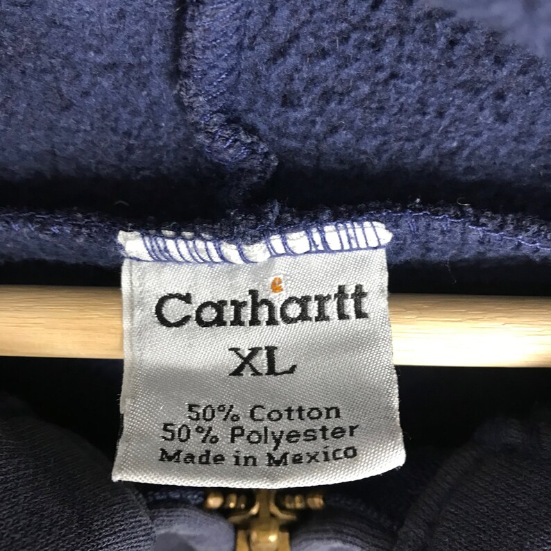 K&ouml;nnte beinhalten: Nahaufnahme des Etiketts eines marineblauen Carhartt-Hoodies. Das wei&szlig;e Etikett zeigt schwarzen Text, darunter "Carhartt XL", Stoffdetails (50 % Baumwolle, 50 % Polyester) und "Made in Mexico".