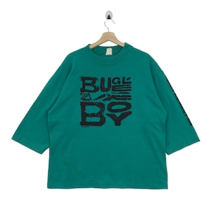 May include: A teal green long-sleeved shirt with a black graphic print that says "BUGLE BOY" in a stylized font.