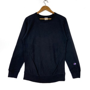 Puede incluir: Una sudadera negra de cuello redondo con mangas largas, colgada de una percha de madera. El logotipo de Champion es visible en la manga izquierda. La sudadera está hecha de un material suave y cómodo.