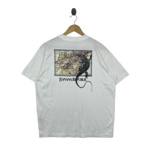 May include: A white short-sleeve t-shirt featuring a graphic on the back. The design includes a rectangular vintage-style map illustration with a black lizard crawling across it. The text BANANA REPUBLIC is printed in black below the graphic.