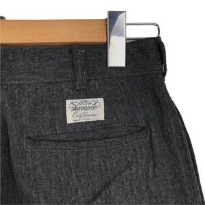 K&ouml;nnte beinhalten: Nahaufnahme einer grauen Hose mit Fischgr&auml;tenmuster, die an einem Holzb&uuml;gel h&auml;ngt. Die Hose hat eine Ges&auml;&szlig;tasche und ein Etikett mit der Aufschrift "DAILY DELIVERY Only Genuine". Der Stoff scheint ein mittelschweres Material zu sein.