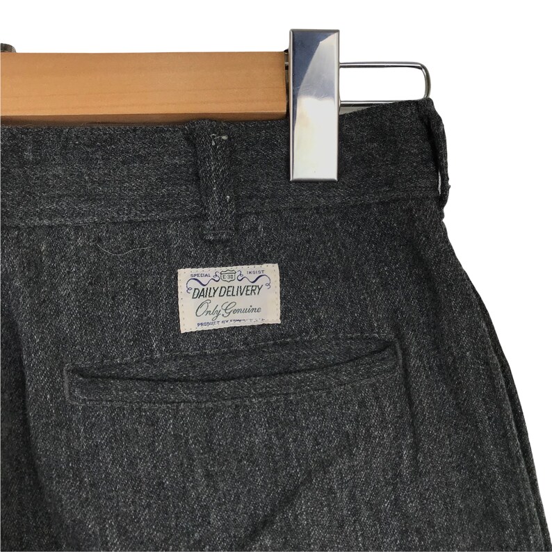 K&ouml;nnte beinhalten: Nahaufnahme einer grauen Hose mit Fischgr&auml;tenmuster, die an einem Holzb&uuml;gel h&auml;ngt. Die Hose hat eine Ges&auml;&szlig;tasche und ein Etikett mit der Aufschrift "DAILY DELIVERY Only Genuine". Der Stoff scheint ein mittelschweres Material zu sein.