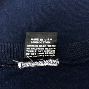 K&ouml;nnte beinhalten: Ein schwarzes Pflegeetikett mit wei&szlig;em Text, der "MADE IN U.S.A 100%COTTON MACHINE WASH WARM NO CHLORINE BLEACH TUMBLE DRY MEDIUM DO NOT IRON DESIGN JUN Planning Co., INC." lautet.