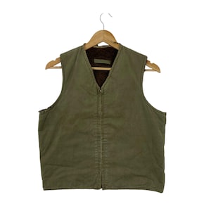May include: Olive green vest with a brown, fuzzy interior, hanging on a wooden hanger. The vest has a V-neck and a full-length zip. The exterior is a solid, muted green colour.