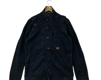 May include: A dark navy blue utility jacket featuring a high collar, a front button placket, and shoulder epaulets. The jacket includes two chest pockets and a small orange G-Star Raw brand label on the left chest area, hanging on a wooden hanger.