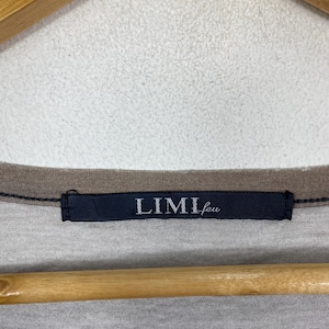 Puede incluir: Una camiseta gris con un cuello acanalado de color marr&oacute;n oscuro y una etiqueta de tela rectangular azul marino cosida en el centro del cuello. La etiqueta muestra el texto blanco LIMI feu en una fuente con serifa, centrado en la prenda.