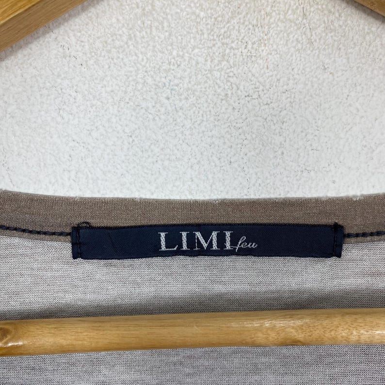 Puede incluir: Una camiseta gris con un cuello acanalado de color marr&oacute;n oscuro y una etiqueta de tela rectangular azul marino cosida en el centro del cuello. La etiqueta muestra el texto blanco LIMI feu en una fuente con serifa, centrado en la prenda.