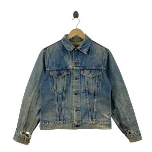 Könnte beinhalten: Eine hellblaue Jeansjacke mit Used-Look, ausgestattet mit einem spitzen Kragen, einer Knopfleiste vorne, zwei Brusttaschen mit geknöpften Klappen sowie ausgefransten, abgenutzten Ärmelbündchen an den langen Ärmeln, präsentiert auf einem Holzkleiderbügel.