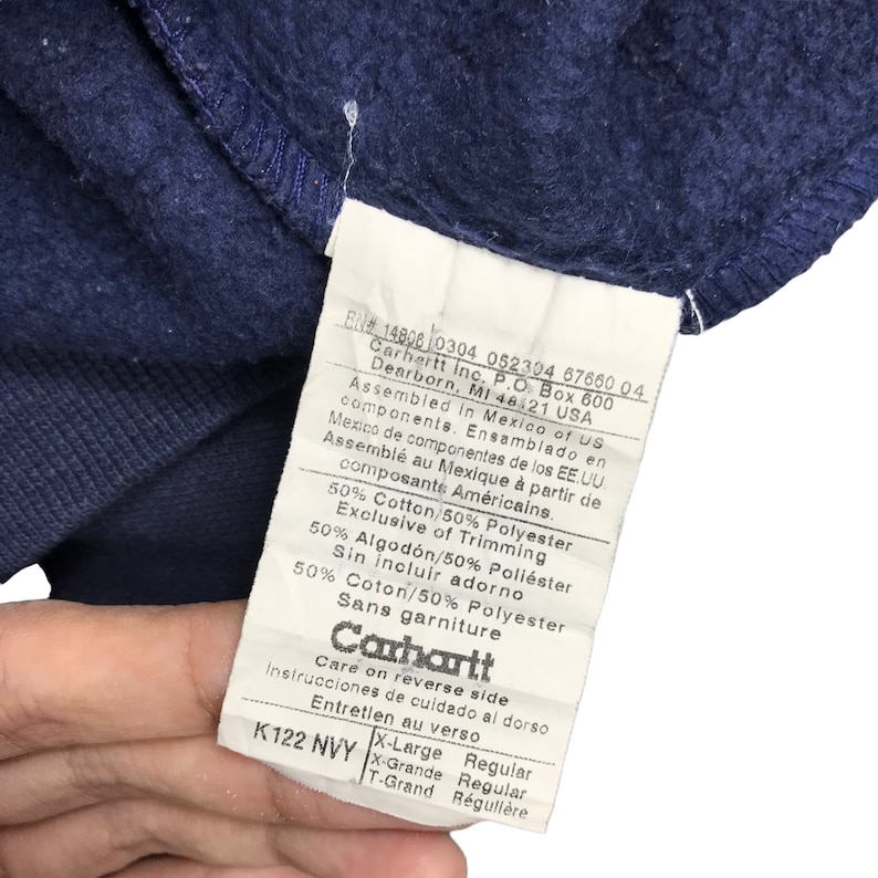 K&ouml;nnte beinhalten: Nahaufnahme eines marineblauen Kleidungsst&uuml;cketiketts. Das Etikett enth&auml;lt das Carhartt-Logo und Details zur Zusammensetzung des Kleidungsst&uuml;cks: 50 % Baumwolle, 50 % Polyester. Das Etikett gibt auch die Montage in Mexiko an.