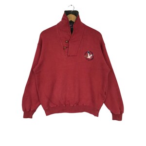 May include: A red sweatshirt with a high collar and two buttons. The collar is lined with green fabric. The sweatshirt features an embroidered logo with a flying duck and the text "CHAPS RALPH LAUREN 1978".