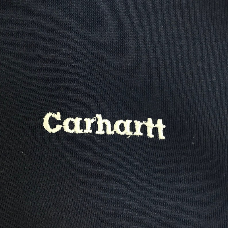 K&ouml;nnte beinhalten: Nahaufnahme eines marineblauen Stoffes mit dem gestickten Wort "Carhartt" in cremefarbenem Garn. Der Text ist zentriert und leicht unterhalb der Mitte des Bildes. Der Stoff hat eine subtile Rippenstruktur.
