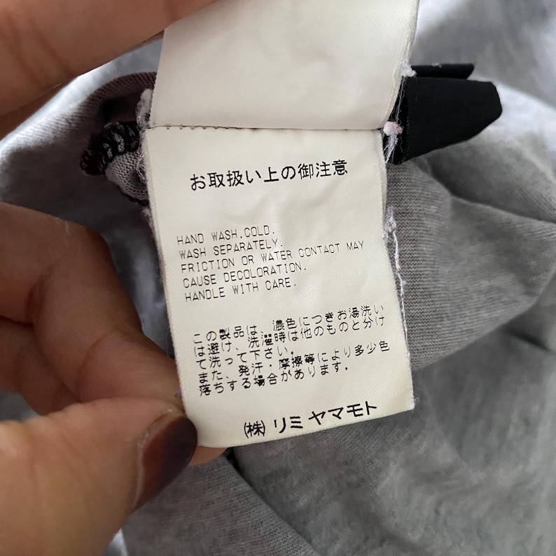 Puede incluir: Una etiqueta de cuidado rectangular blanca unida a una prenda gris. La etiqueta presenta texto en negro con las instrucciones: HAND WASH COLD, WASH SEPARATELY, FRICTION OR WATER CONTACT MAY CAUSE DECOLORATION, HANDLE WITH CARE, y texto en japon&eacute;s.