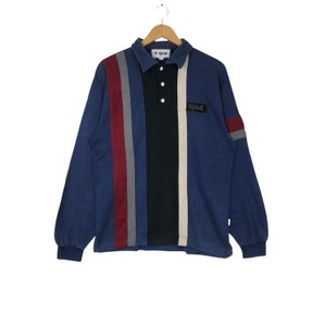 Peut inclure: Un polo bleu à manches longues avec des rayures verticales rouges, grises, noires et blanc cassé. Le polo a un col, trois boutons et un petit écusson avec du texte sur la poitrine. Le polo est suspendu à un cintre en bois.