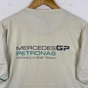 May include: A beige short-sleeved t-shirt featuring the Mercedes GP Petronas Formula One Team logo printed in black and teal on the chest. The shirt has a crew neckline and a semi-circular stitched panel detail below the collar.