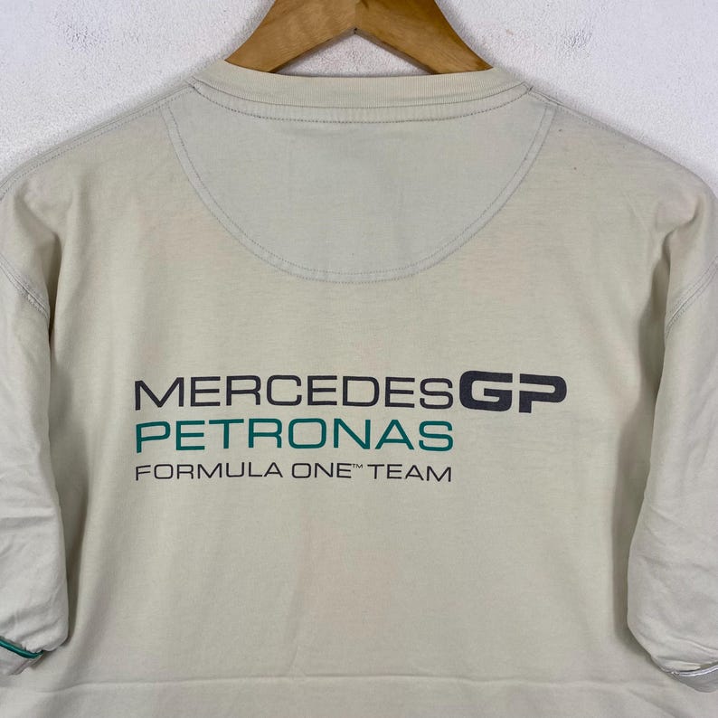 May include: A beige short-sleeved t-shirt featuring the Mercedes GP Petronas Formula One Team logo printed in black and teal on the chest. The shirt has a crew neckline and a semi-circular stitched panel detail below the collar.