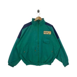 May include: A vintage green jacket with navy blue shoulder accents. The jacket features a full zipper, two front pockets, and the text "Benetton Formula 1" on a patch. The jacket is hanging on a wooden hanger.