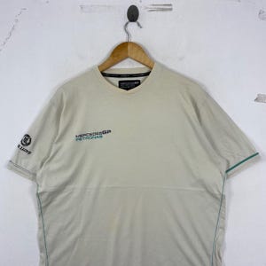 May include: A beige short-sleeved t-shirt featuring the Mercedes GP Petronas logo on the chest in black and teal. The right sleeve displays a black logo with the text Henri Lloyd. The shirt includes thin teal piping along the side seams.