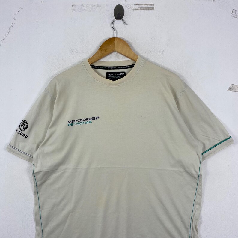 May include: A beige short-sleeved t-shirt featuring the Mercedes GP Petronas logo on the chest in black and teal. The right sleeve displays a black logo with the text Henri Lloyd. The shirt includes thin teal piping along the side seams.