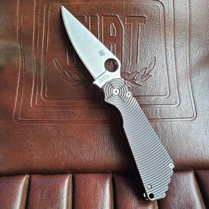 May include: A silver folding knife with a ridged handle and a pointed blade. The knife is resting on a brown leather surface with the embossed word "FIAT". The blade has the text "CPM S30V".