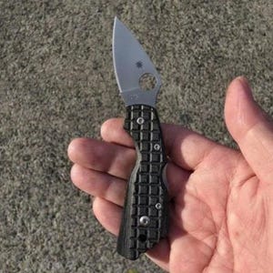 May include: A folding pocket knife with a silver-coloured metal blade featuring a circular cutout near the base and a black textured handle with a grid pattern, secured by visible silver screws along the side of the handle grip.