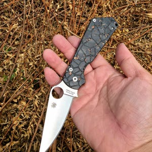 May include: A folding knife with a silver blade and a black and copper patterned handle. The blade has a circular cutout near the handle and the brand name "Spyderco" is visible. The handle has a geometric design.