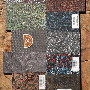 Puede incluir: Una colección de cachas rectangulares para mangos de cuchillos con varios patrones y colores, incluyendo rojo, verde, azul y negro. Una cacha presenta un tejido de fibra de carbono y un logotipo de "Knife Handle Supply". Las cachas están dispuestas sobre una superficie de madera.