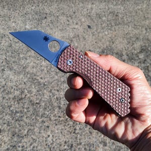 May include: A pocket knife with a brown textured handle and a silver blade. The blade has a unique shape and a circular hole near the handle. The knife is held in a hand against a grey background. The brand name "Spyderco" is visible on the blade.