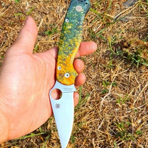 May include: A folding knife with a blade and a handle. The handle is a mix of green, yellow, and orange. The blade is silver and has the text "CPM S30V" and a spider logo. The knife is held in a hand.