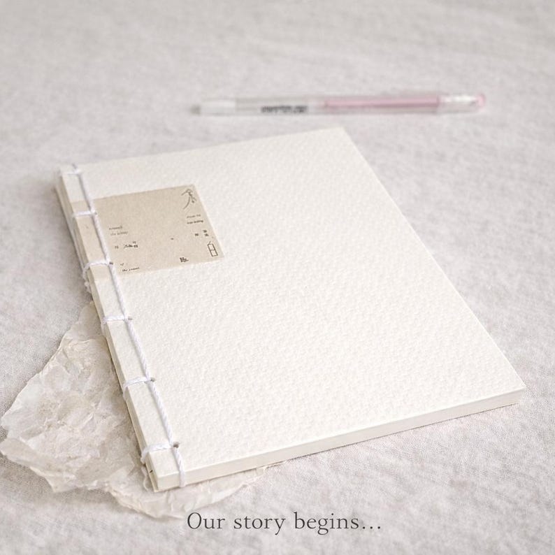 Pu&ograve; includere: Un diario bianco strutturato con rilegatura cucita e una piccola etichetta. Una penna rosa &egrave; sullo sfondo. La frase "Our story begins..." &egrave; stampata in basso nell'immagine.