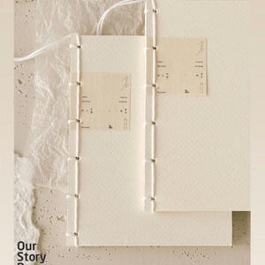 Pu&ograve; includere: Due quaderni cuciti a mano color bianco sporco con pagine color crema. I quaderni sono rilegati con filo bianco e presentano piccole etichette rettangolari con testo e illustrazioni minimaliste. La frase "Our Story Begins" &egrave; stampata in basso a sinistra.