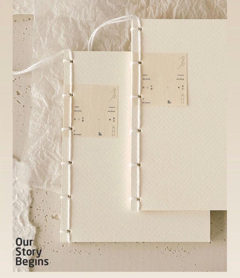 Pu&ograve; includere: Due quaderni cuciti a mano color bianco sporco con pagine color crema. I quaderni sono rilegati con filo bianco e presentano piccole etichette rettangolari con testo e illustrazioni minimaliste. La frase "Our Story Begins" &egrave; stampata in basso a sinistra.