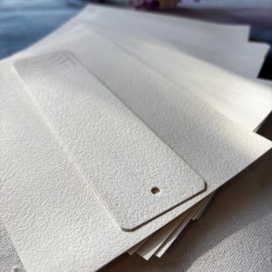 以下が含まれることがあります： 質感のあるオフホワイトの長方形の紙製ブックマークの束。各ブックマークは角が丸くなっており、下部中央に小さな円形の穴が開けられ、上端には一連のエンボス加工された曲線が施されています。