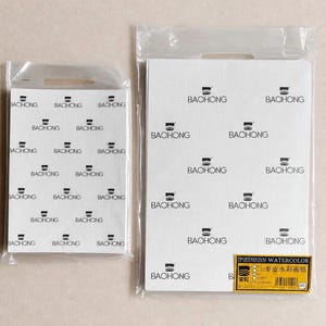 Könnte beinhalten: Zwei rechteckige Blöcke aus weißem Aquarellpapier, jeweils in einer durchsichtigen Plastiktüte mit Tragegriff verpackt. Das Papier zeigt ein sich wiederholendes schwarzes Logo-Muster mit der Aufschrift BACHONG. Ein gelbes Etikett auf dem größeren Block trägt den Text PROFESSIONAL WATERCOLOR.