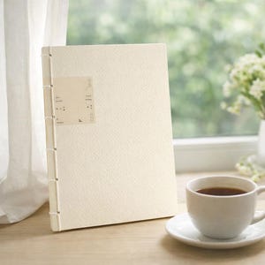May include: A cream-coloured journal with a textured cover and stitched binding. A small rectangular label with text is affixed to the front. A white teacup and saucer sit in the foreground, with a window and greenery in the background.